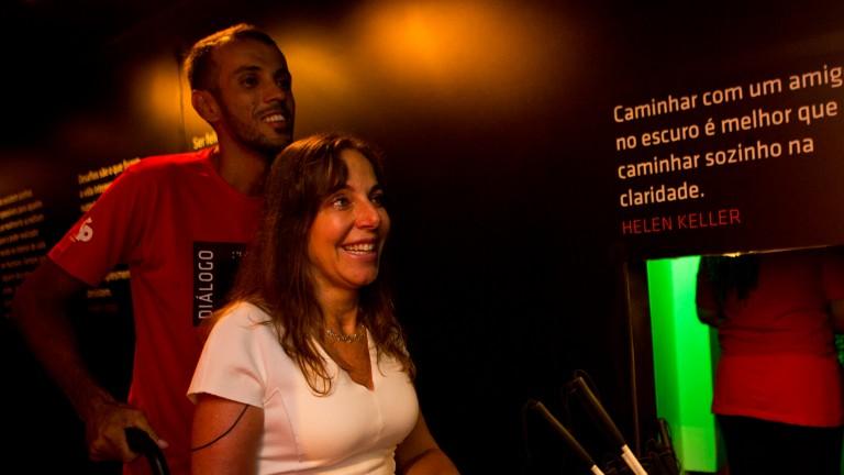 Foto da Senadora Mara Gabrilli, sorridente na sua cadeira de rodas, sendo levada por um dos guias da Exposição, após sua visita. Ao fundo a famosa frase de Helen Keller, na parede: "Caminhar com um amigo noescuro é melhor que caminhar sozinho na claridade".