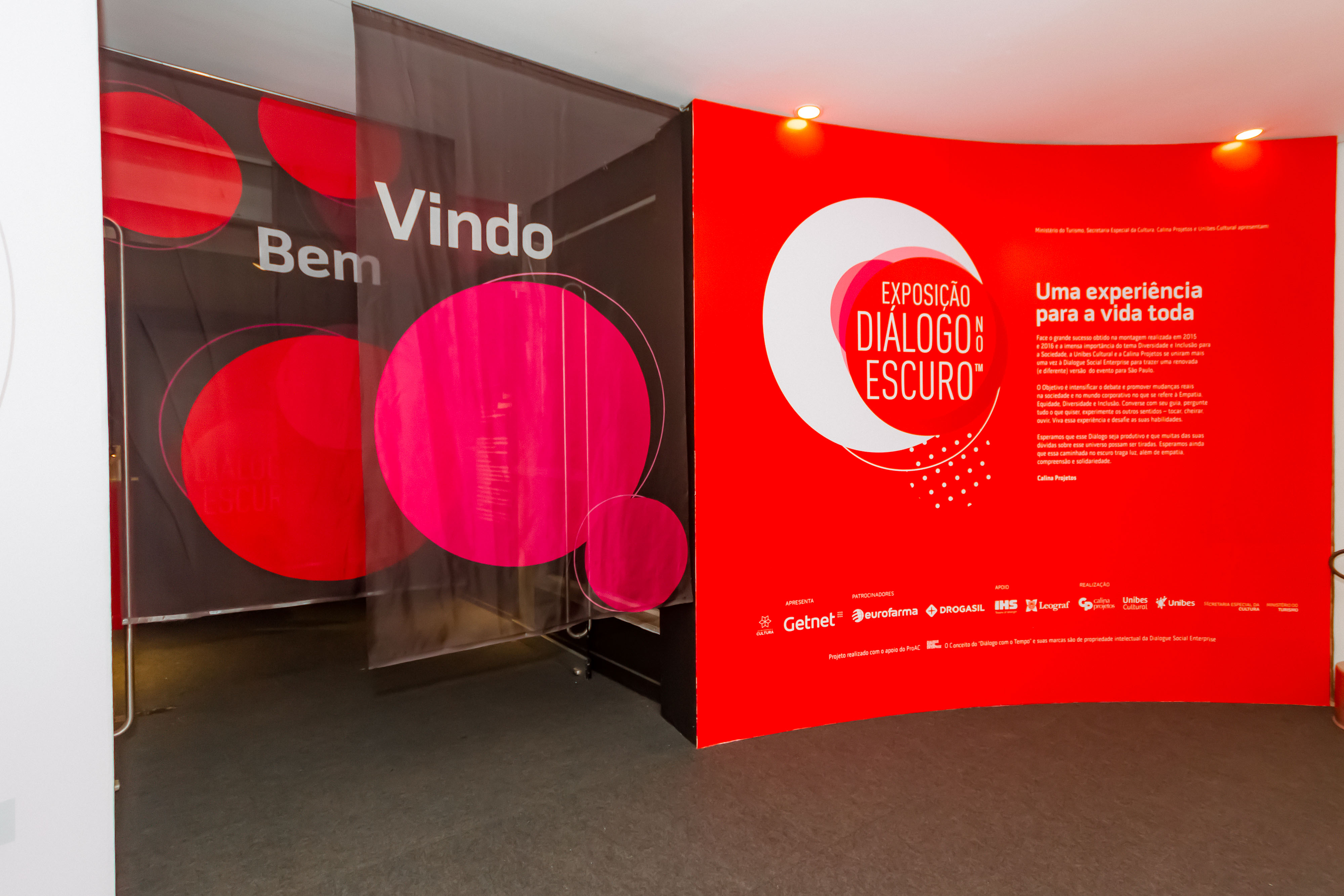Foto da entrada com cortinas escritas bem vindo e parede vermelha a direita com logo da exposição e textos em branco