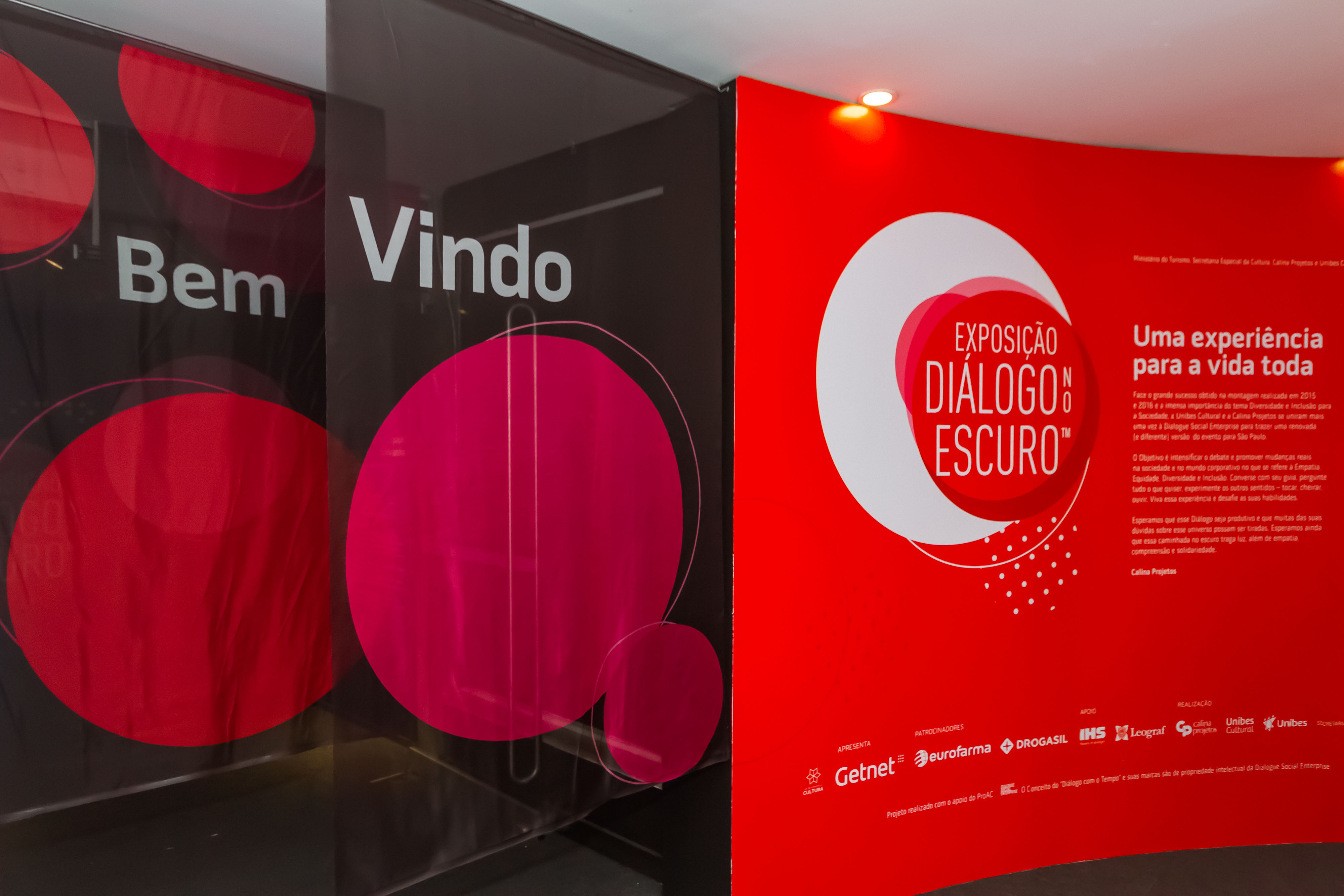 Foto da entrada com cortinas escritas bem vindo e parede vermelha a direita com logo da exposição e textos em branco