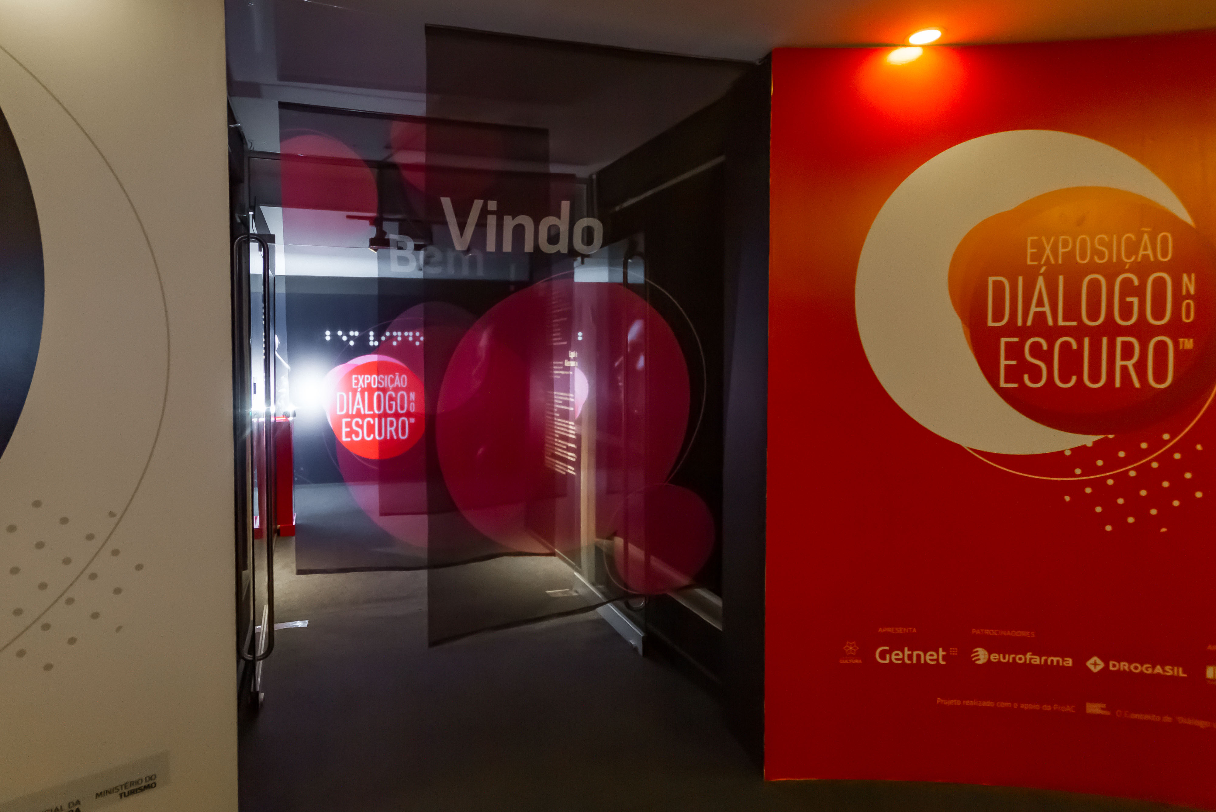 Foto da entrada com cortinas escritas bem vindo, logo da exposição ao fundo além da cortina e parede vermelha a direita com logo da exposição